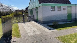 Een klein, laag gebouw met een groene deur en luiken, nummer 226, naast een haag en een verhard pad.