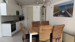 A modern kitchen with white cabinets, a stove, and a microwave. There is a dining table with wicker chairs and a painting of a beach scene on the wall. - 3 bedroom accommodation in Julianadorp (Julianadorp)