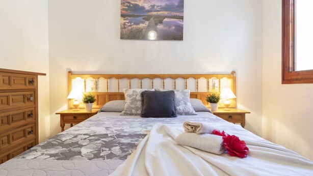 Ein Schlafzimmer mit einem Holzkopfteil, zwei Nachttischen mit Lampen, einer blumigen Bettdecke und einem Landschaftsbild an der Wand.