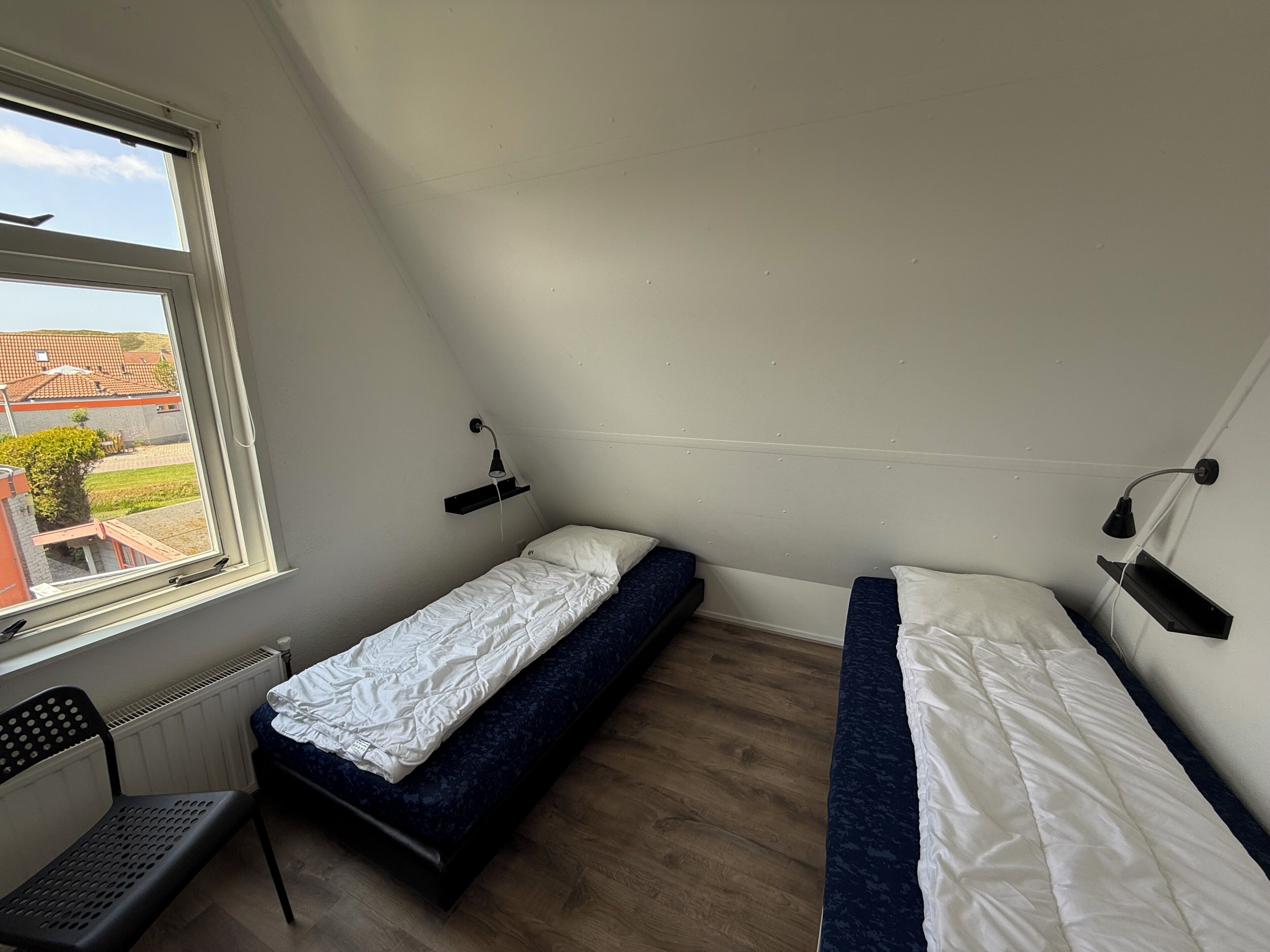 Eine kleine, einfache Wohnung mit zwei Betten, einem Schreibtisch und einem Fenster mit Blick auf eine Wohngegend.