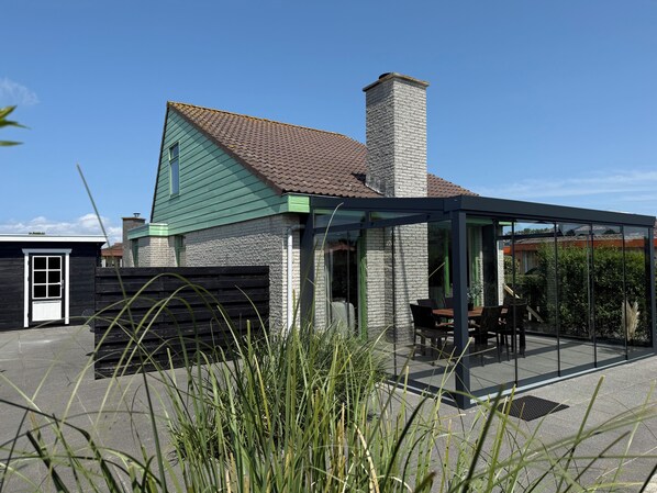 A house with a green roof, a stone chimney, and a glass-enclosed patio area with outdoor furniture. - 3 bedroom accommodation in Julianadorp (Julianadorp)