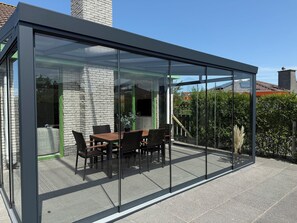 A modern glass-enclosed patio with a dining table and chairs, surrounded by a stone wall and greenery. - 3 bedroom accommodation in Julianadorp (Julianadorp)