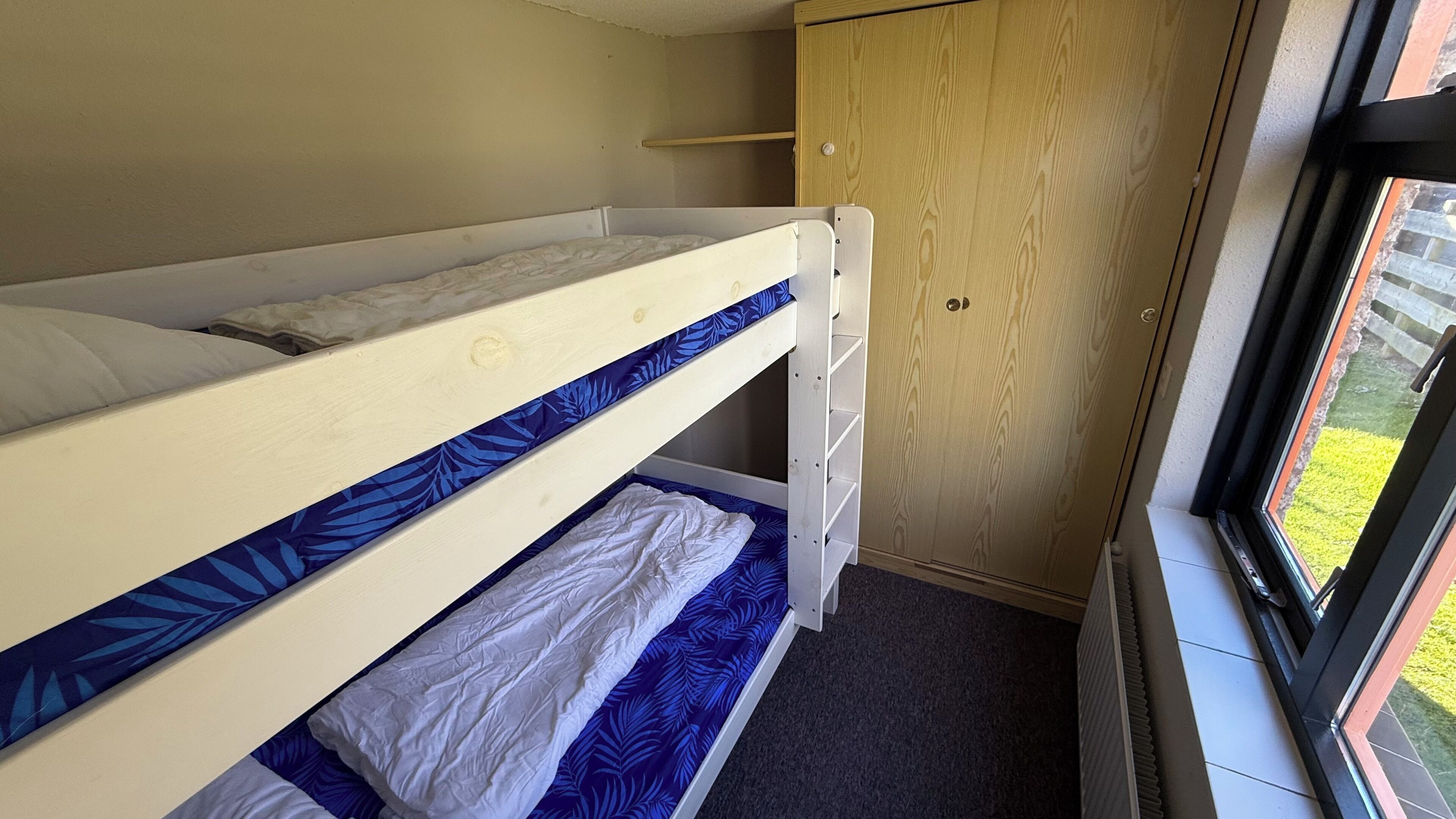 Een stapelbed met een onderbed, een houten garderobe en een raam met uitzicht op groen.