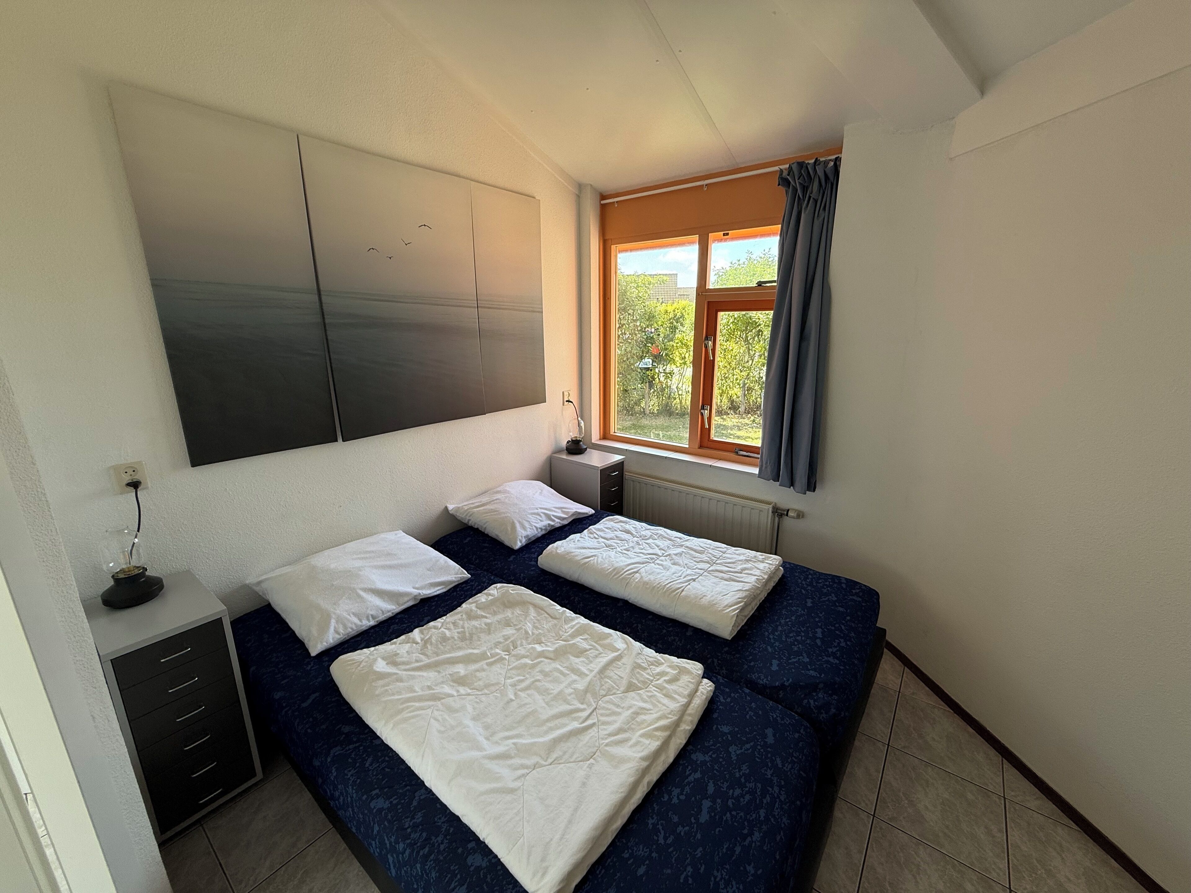 Una camera da letto con due letti, una grande finestra con tende, un comodino con una lampada e una TV a muro.