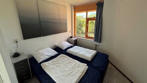 Un dormitorio con dos camas, un ventanal con cortinas, una mesita de noche con lámpara y un televisor de pared.