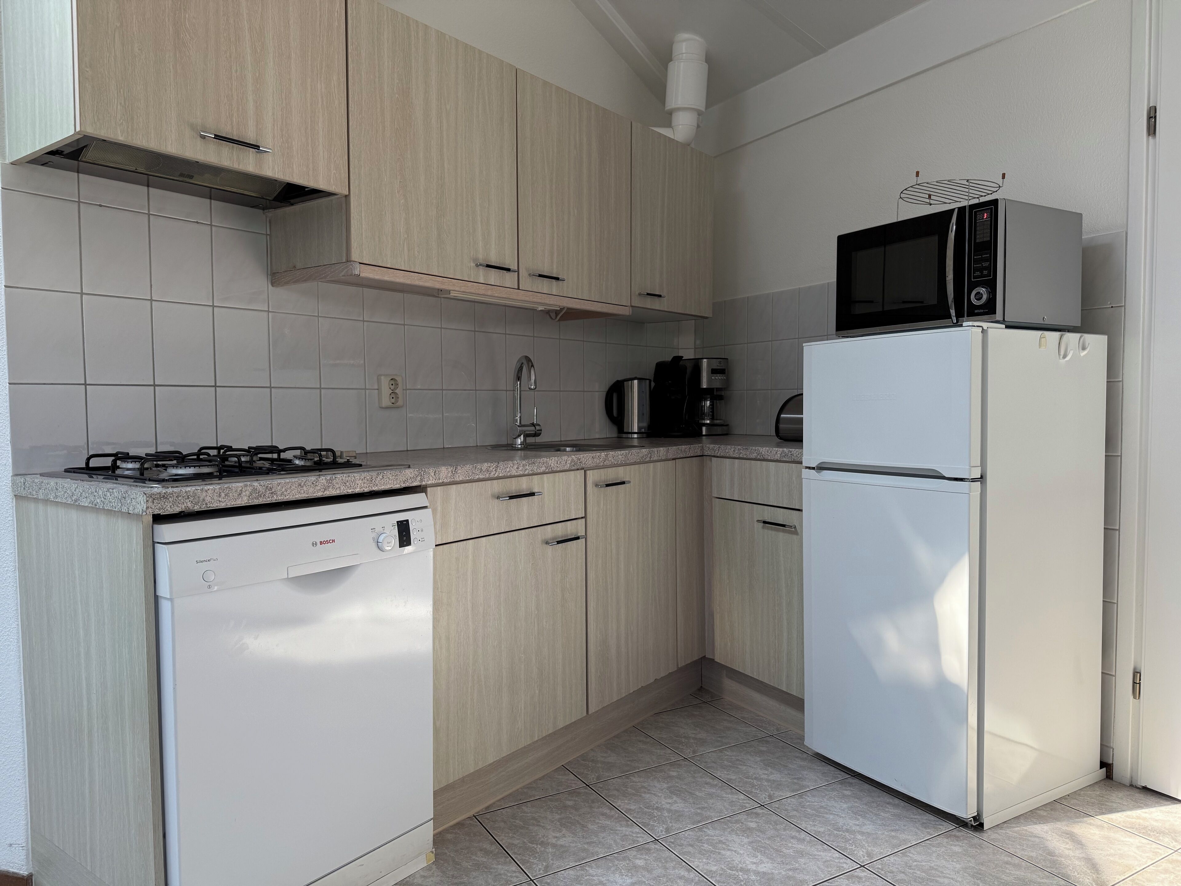 Ett modernt kök med ljusa träskåp, en diskmaskin från Bosch, ett vitt kylskåp, en mikrovågsugn och en gasspis.