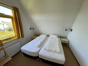 A small, single-room bedroom with two beds, a window with curtains, a radiator, and a chair.