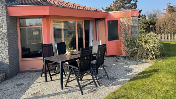 A small, single-story building with a red exterior and large windows. It features a patio with a table and chairs, surrounded by a gravel area and a grassy lawn.