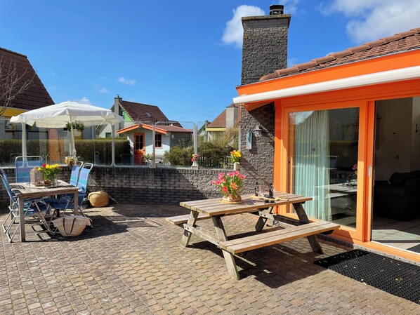 A patio area with a wooden table and chairs, a stone wall, and a glass door leading to a room.