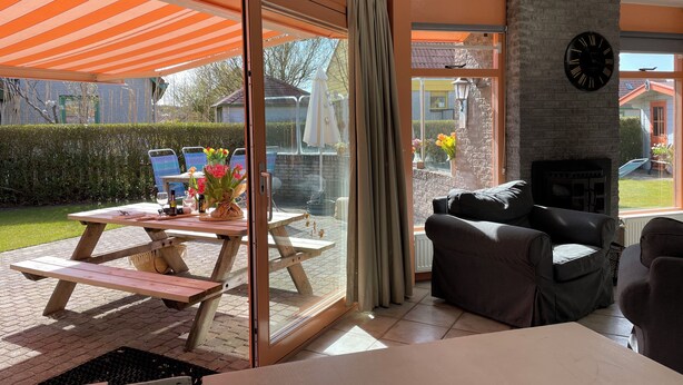 A modern living room with a fireplace, a dining area with a table and chairs, and a large sliding glass door leading to a garden. - 2 bedroom accommodation in Julianadorp aan Zee (Julianadorp aan Zee)