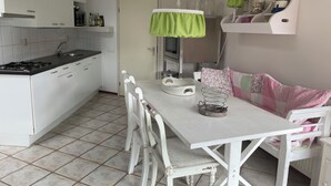 A kitchen with white cabinets, a stove, and a dining table with chairs. There is a hanging light fixture and a shelf with decorative items. - 2 bedroom accommodation in Julianadorp aan Zee (Julianadorp aan Zee)