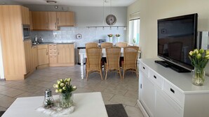 A modern kitchen with wooden cabinets, a dining area with wicker chairs, a flat-screen TV, and a vase with flowers on a white table.