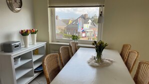 A dining area with a white table, wicker chairs, a vase with flowers, a clock on the wall, and a window with a view of buildings.