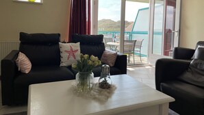 A living room with a black leather sofa, a white coffee table with a vase of flowers, and a view of the outside balcony.