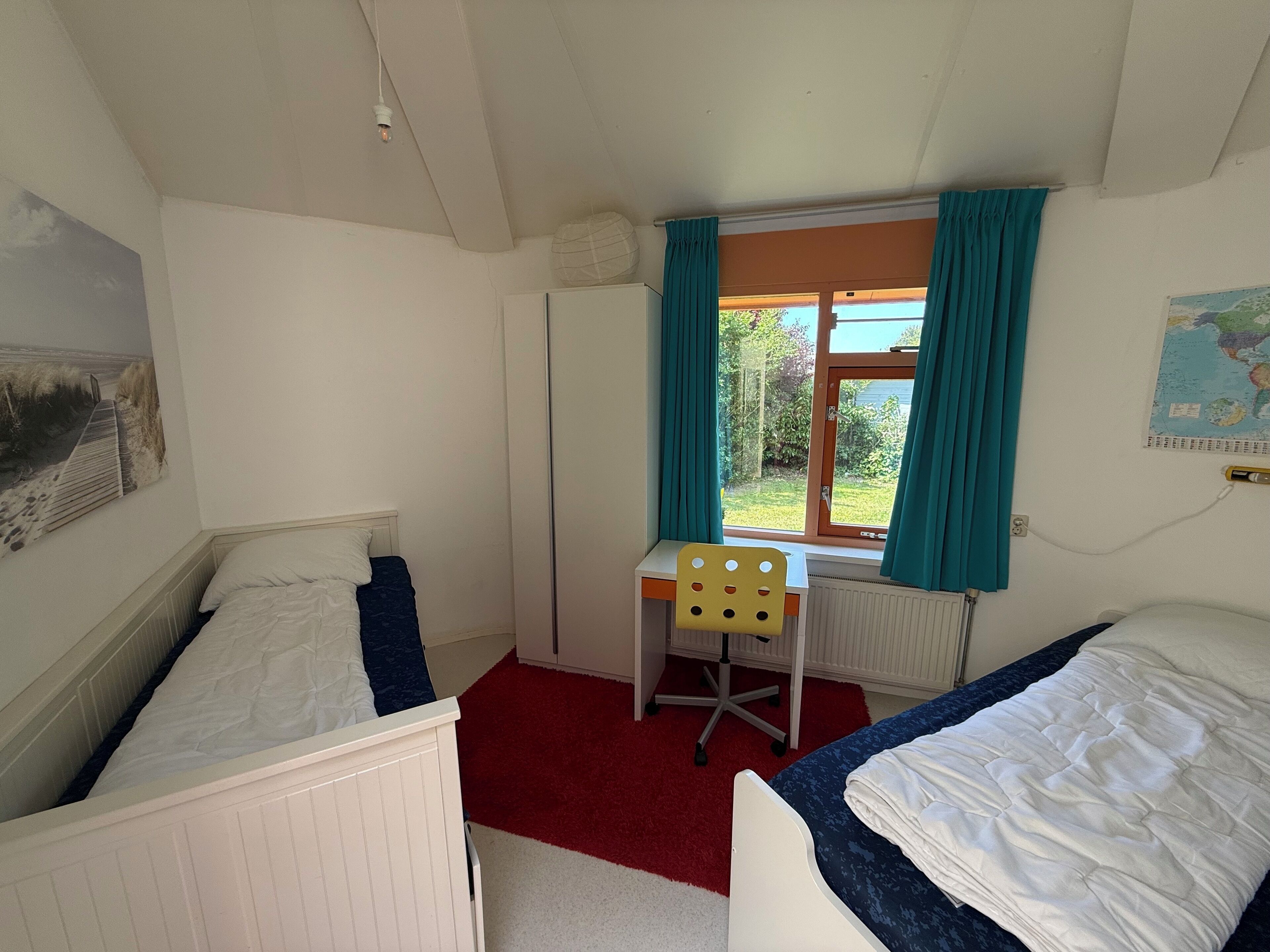A room with two beds, a desk, a chair, a radiator, a window with curtains, and a map on the wall.