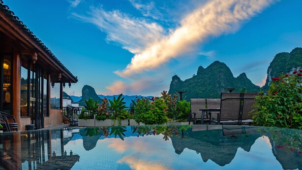 A serene pool area with a clear reflection of the sky and mountains, surrounded by lush greenery and traditional wooden architecture. - Yangshuo Mountain River Ballad Hotel (Guilin)