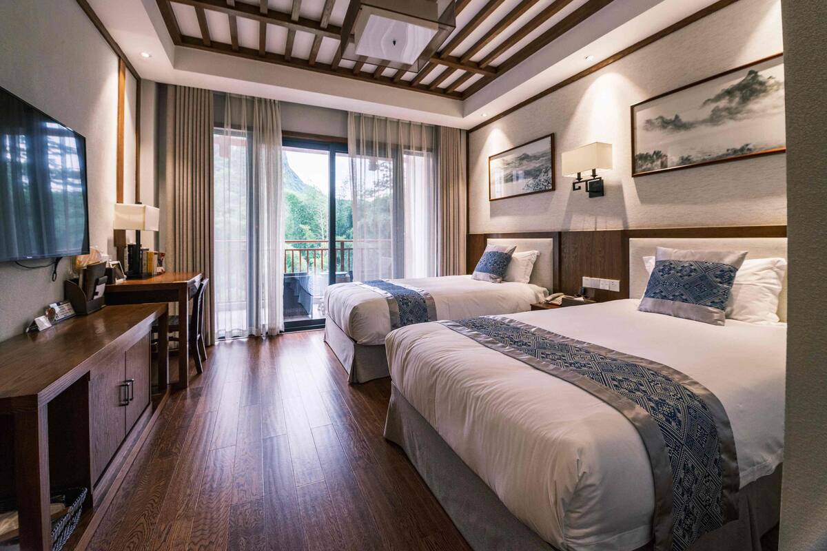A hotel room with two beds, a desk with a lamp, a TV, and a balcony with curtains.