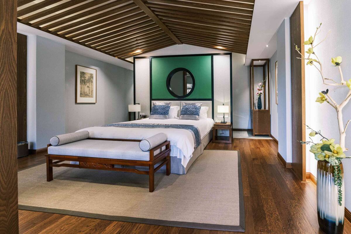 A modern bedroom with a large bed, a wooden ceiling, a green accent wall, and a vase with flowers.