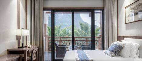 A hotel room with a large bed, a desk, and a view of a mountainous landscape through a sliding glass door.