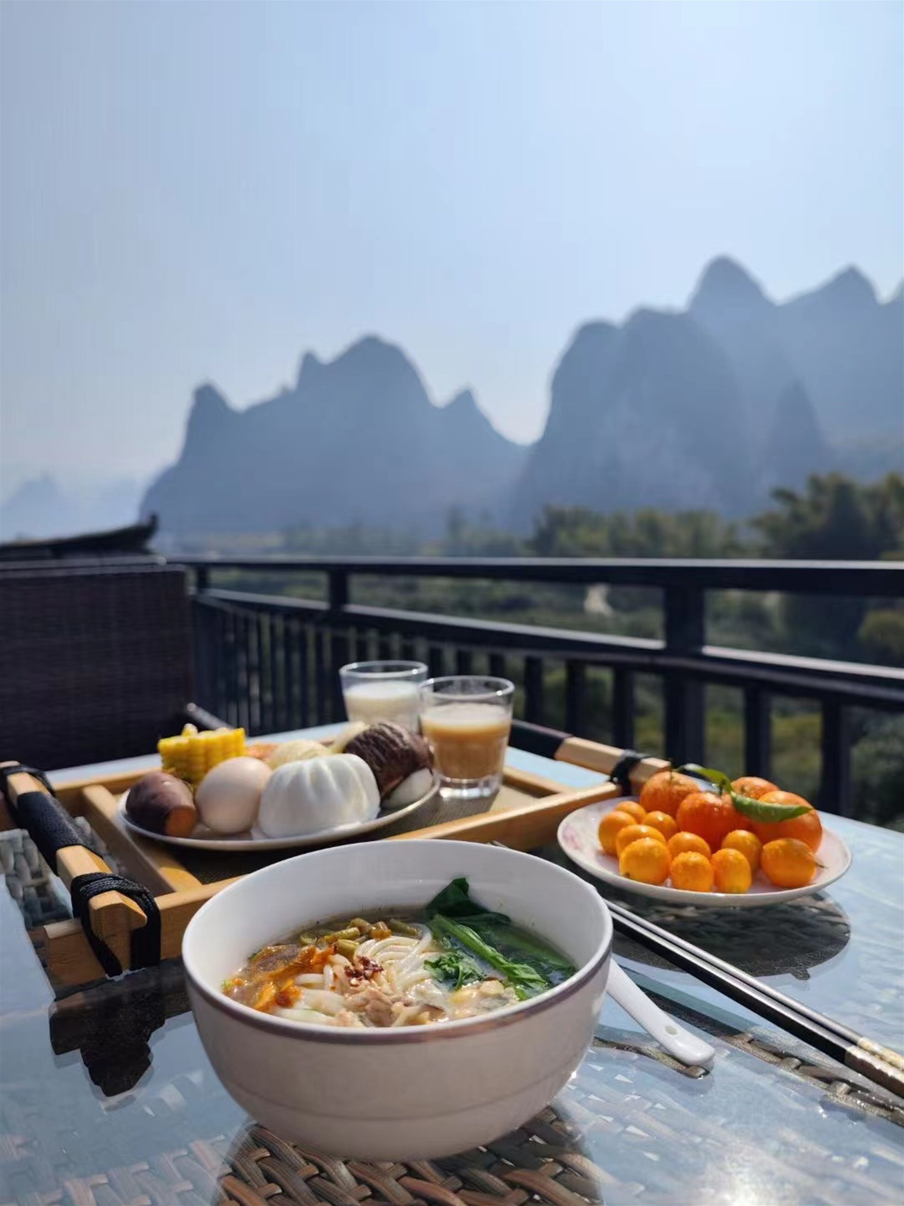 A table with a bowl of soup, a plate of oranges, and a plate of corn on the table, with a mountainous view in the background.