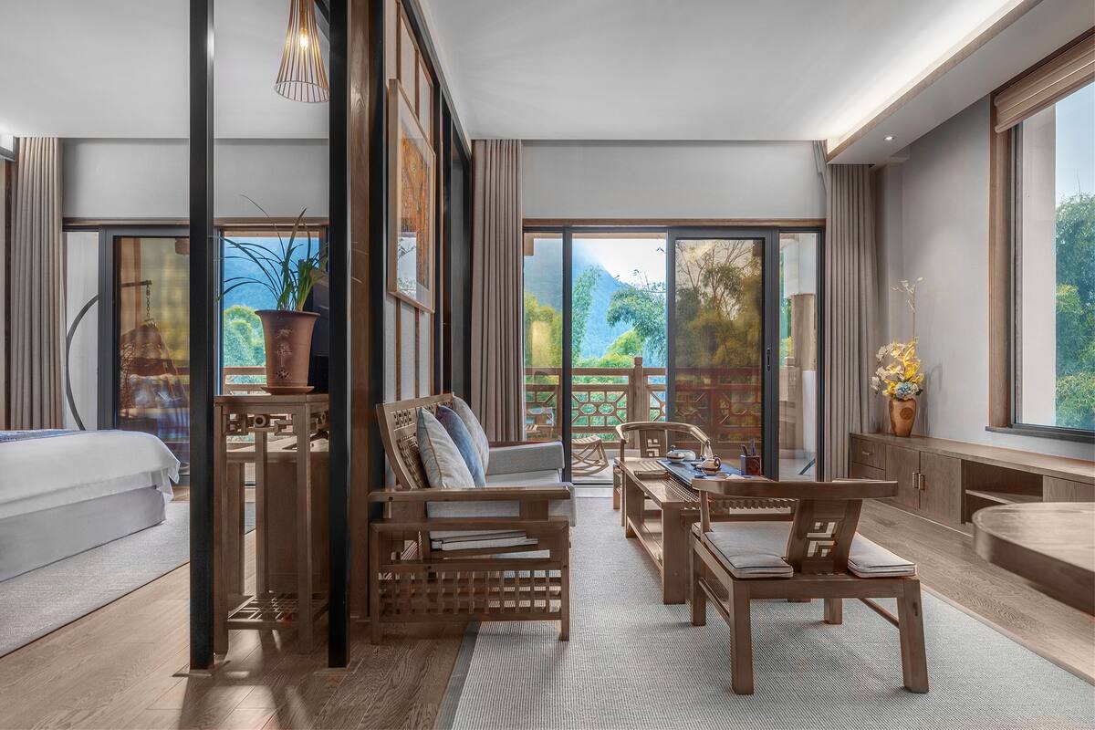 A modern hotel room with a large sliding glass door leading to a balcony, a wooden sofa, a dining table with chairs, and a small kitchen area.