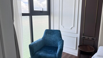 A room with a large window offering a view of the sea, a blue armchair, a wooden side table, and a white door with a panel.