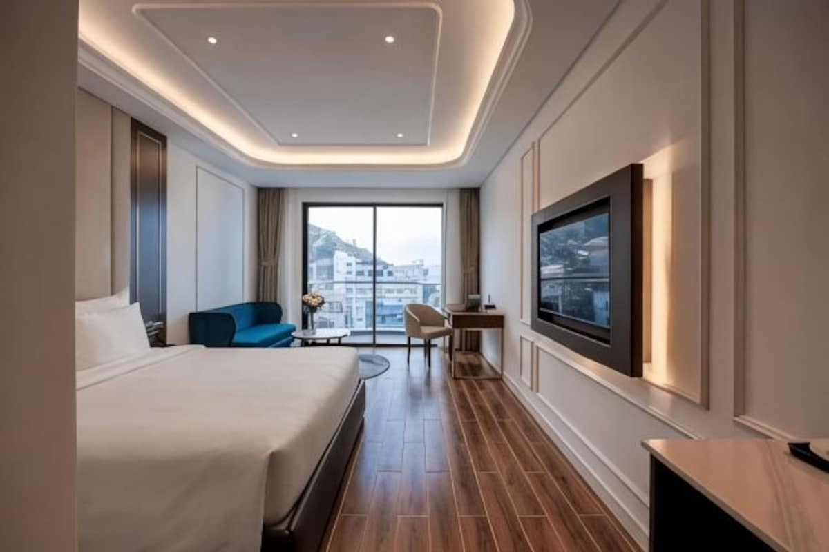 A modern hotel room with a large bed, a seating area with a blue sofa, a small table, a TV mounted on the wall, and a city view through the window.