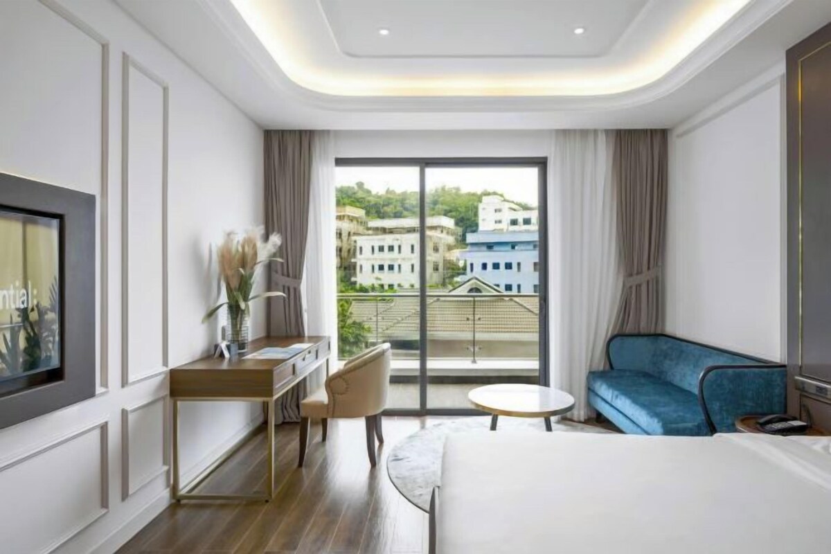 Une chambre d’hôtel moderne dotée d’un grand lit, d’un bureau avec une chaise, d’une table ronde, d’un canapé bleu et d’une grande fenêtre donnant sur des bâtiments et des espaces verts.