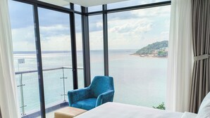 Hotelkamer met een ruim balkon, een bed, een blauwe stoel en uitzicht op zee en een eiland in de verte.