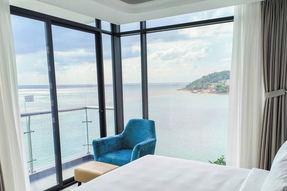 Une chambre d’hôtel avec un grand balcon, un lit, une chaise bleue et une vue sur la mer et une île lointaine.
