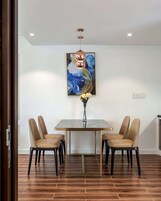 Un coin repas comprenant une table rectangulaire, quatre chaises, un vase avec des fleurs, un luminaire au plafond et un tableau accroché au mur.