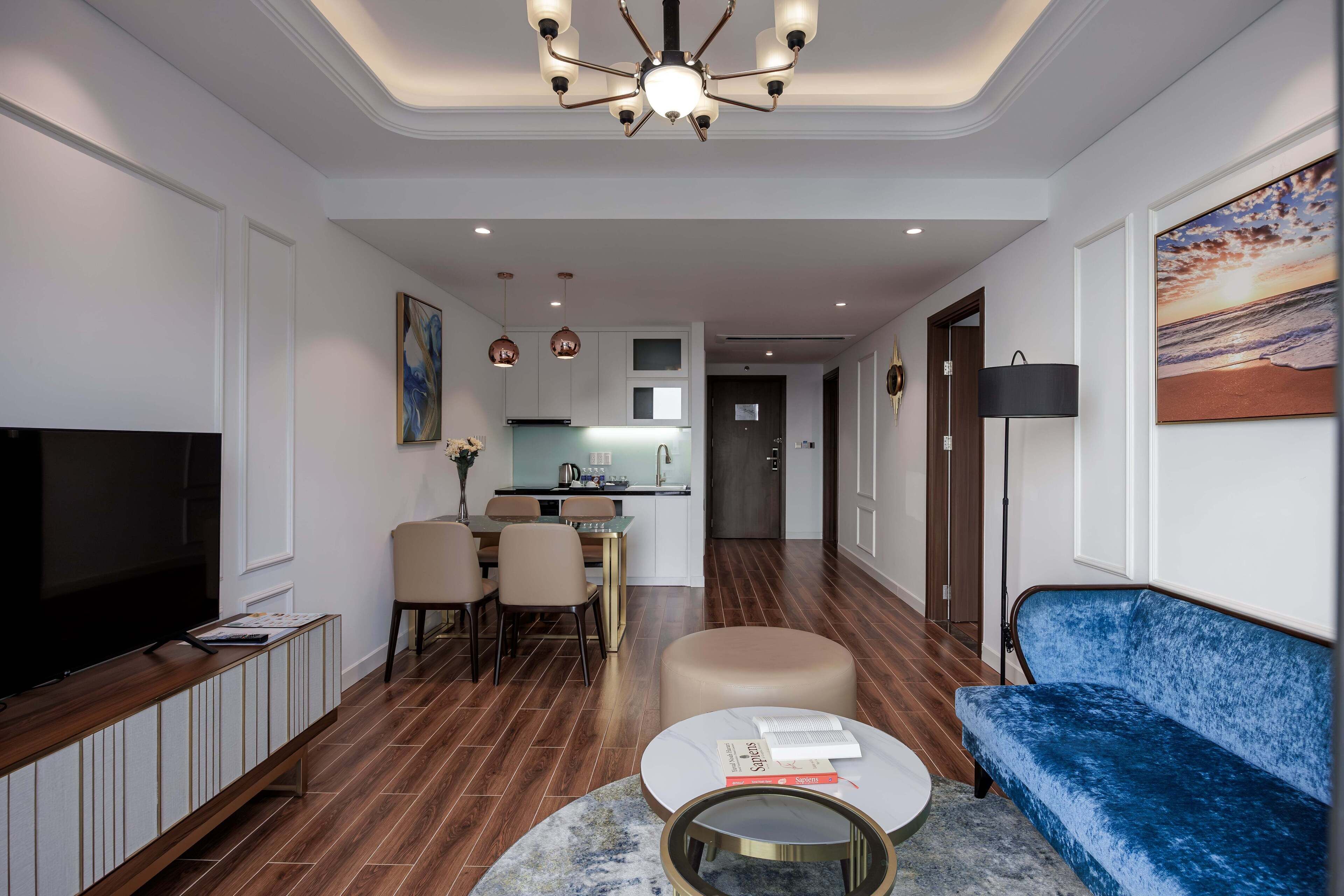 A modern living room with a flat-screen TV, a blue sofa, a round coffee table, and a dining area with chairs and a kitchen in the background.