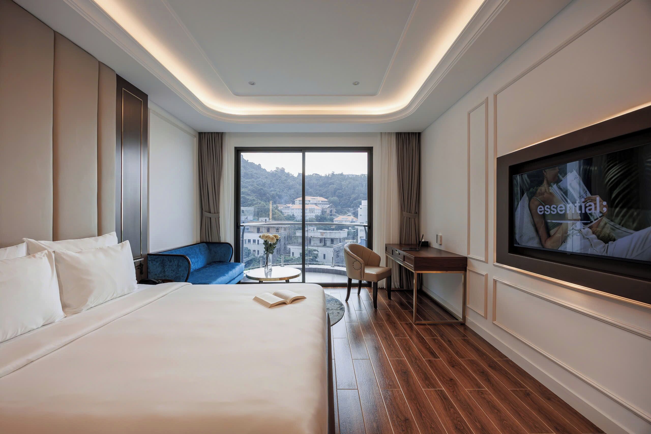 A hotel room with a large bed, a desk, a chair, a TV, and a view of a cityscape through a large window.