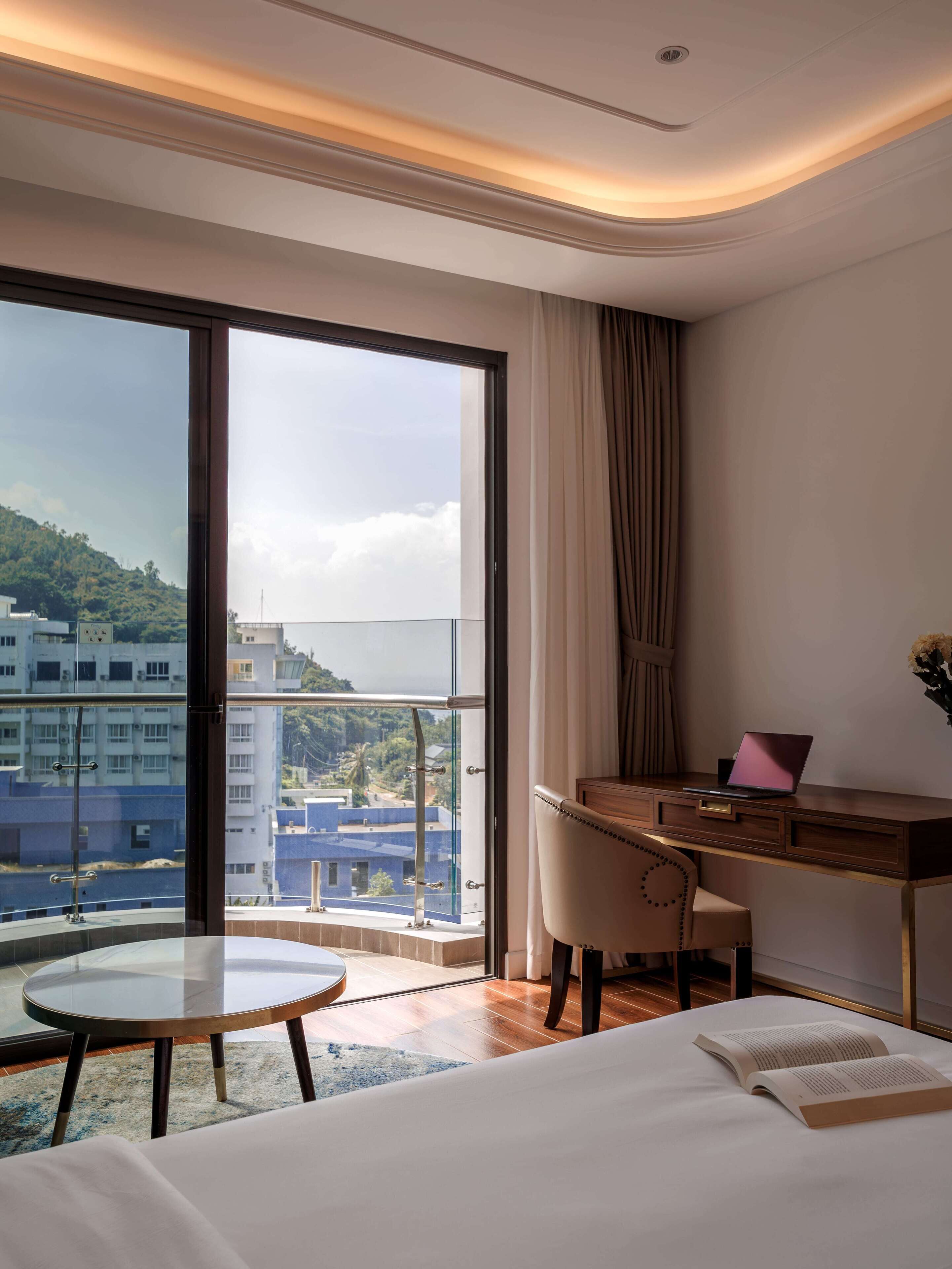 Une chambre d’hôtel avec une grande fenêtre, un bureau, une chaise, une table ronde et un lit.