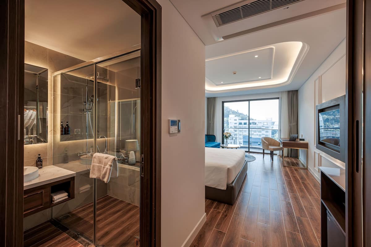 Une chambre d’hôtel moderne dotée d’une douche en verre, d’une salle de bain séparée, d’un lit avec télévision et d’un bureau avec un ordinateur portable.
