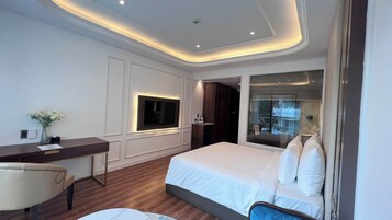 A modern hotel room with a large bed, a desk, a chair, a TV mounted on the wall, and a glass shower enclosure.