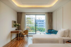 A hotel room with a bed, a desk, a chair, a small table, and a view of buildings through a large window.