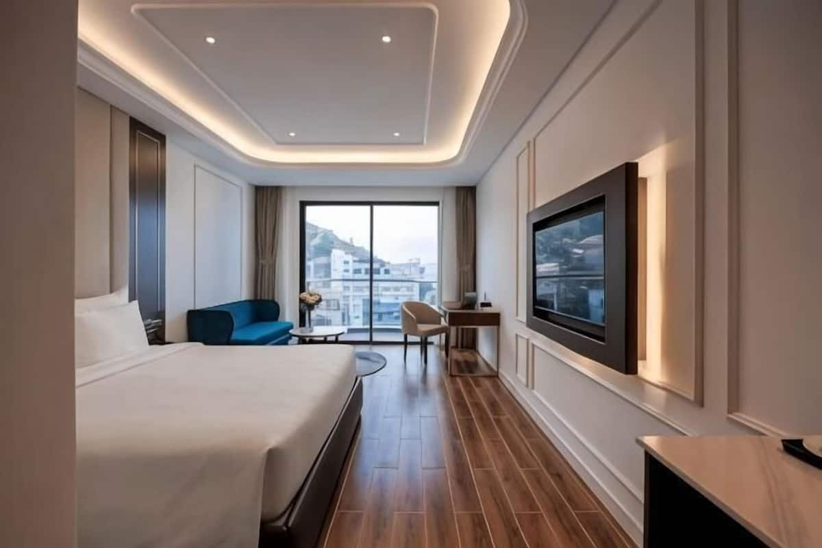 Une chambre d’hôtel moderne avec un grand lit, un coin salon avec un canapé bleu, une petite table, un téléviseur fixé au mur et une vue sur la ville par la fenêtre.