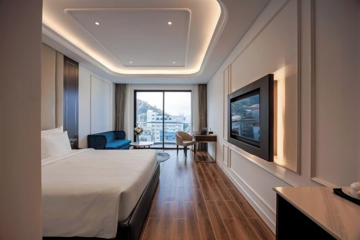 A modern hotel room with a large bed, a desk, a chair, and a TV mounted on the wall.
