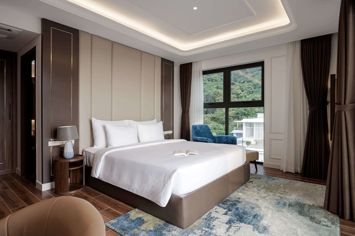Une chambre d’hôtel moderne avec un grand lit, une table de chevet avec une lampe, une chaise bleue et une vue sur la verdure par la fenêtre.
