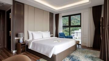 Habitación de hotel moderna con una cama grande, una mesita de noche con una lámpara, una silla azul y vistas a zonas verdes por la ventana.