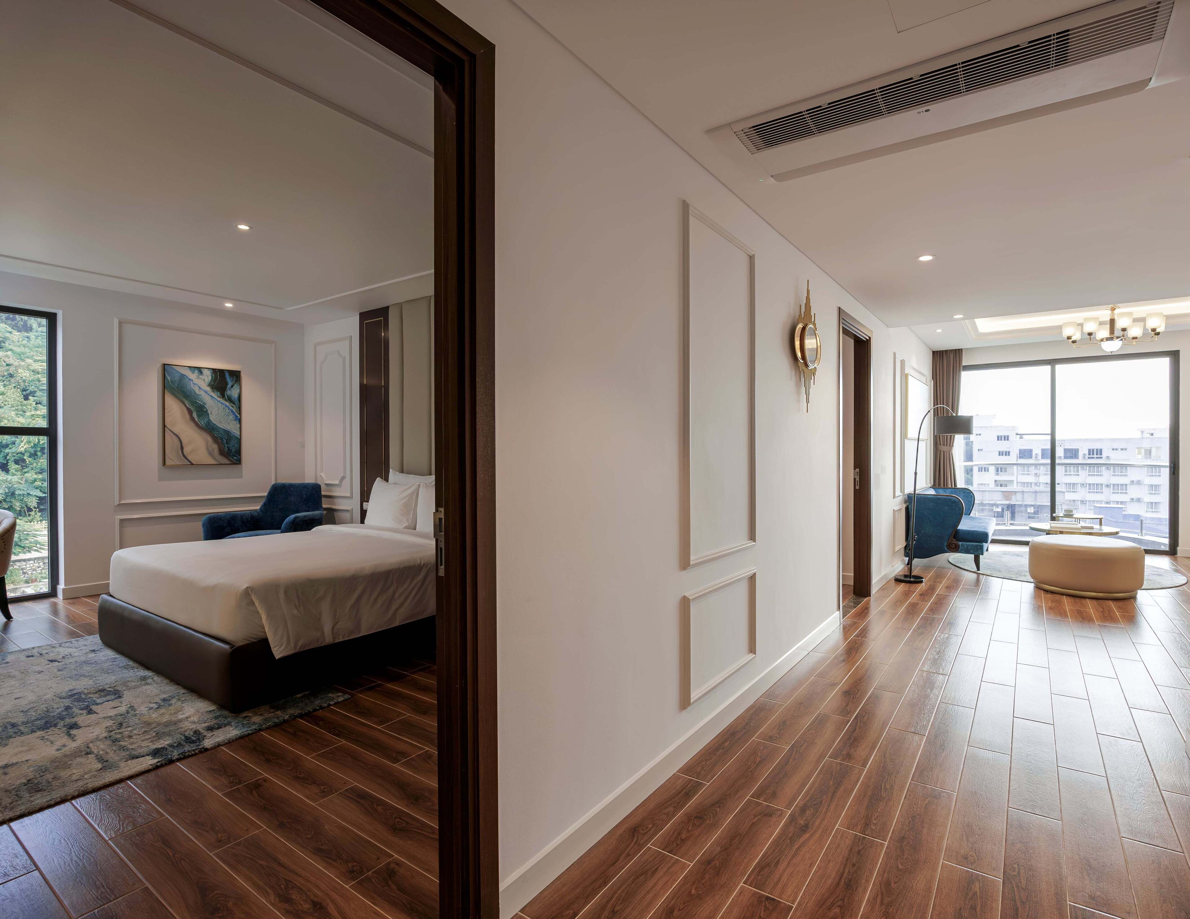 Une chambre d’hôtel moderne dotée d’un grand lit, d’un coin salon avec un canapé et un fauteuil, d’une grande fenêtre offrant une vue sur la ville et d’un couloir menant à une autre chambre.