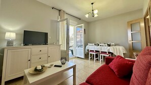 Un espace de vie compact comprenant un téléviseur posé sur un meuble, un coin repas avec une table et des chaises, et un canapé rouge.