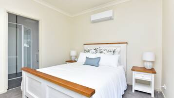 A bedroom with a bed, bedside tables, a nightstand, a wall-mounted air conditioner, and a glass shower enclosure.