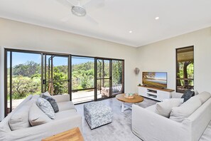 A modern living room with large windows, a ceiling fan, a flat-screen TV, and a wooden coffee table. - Kookaburra Range (North Rothbury)