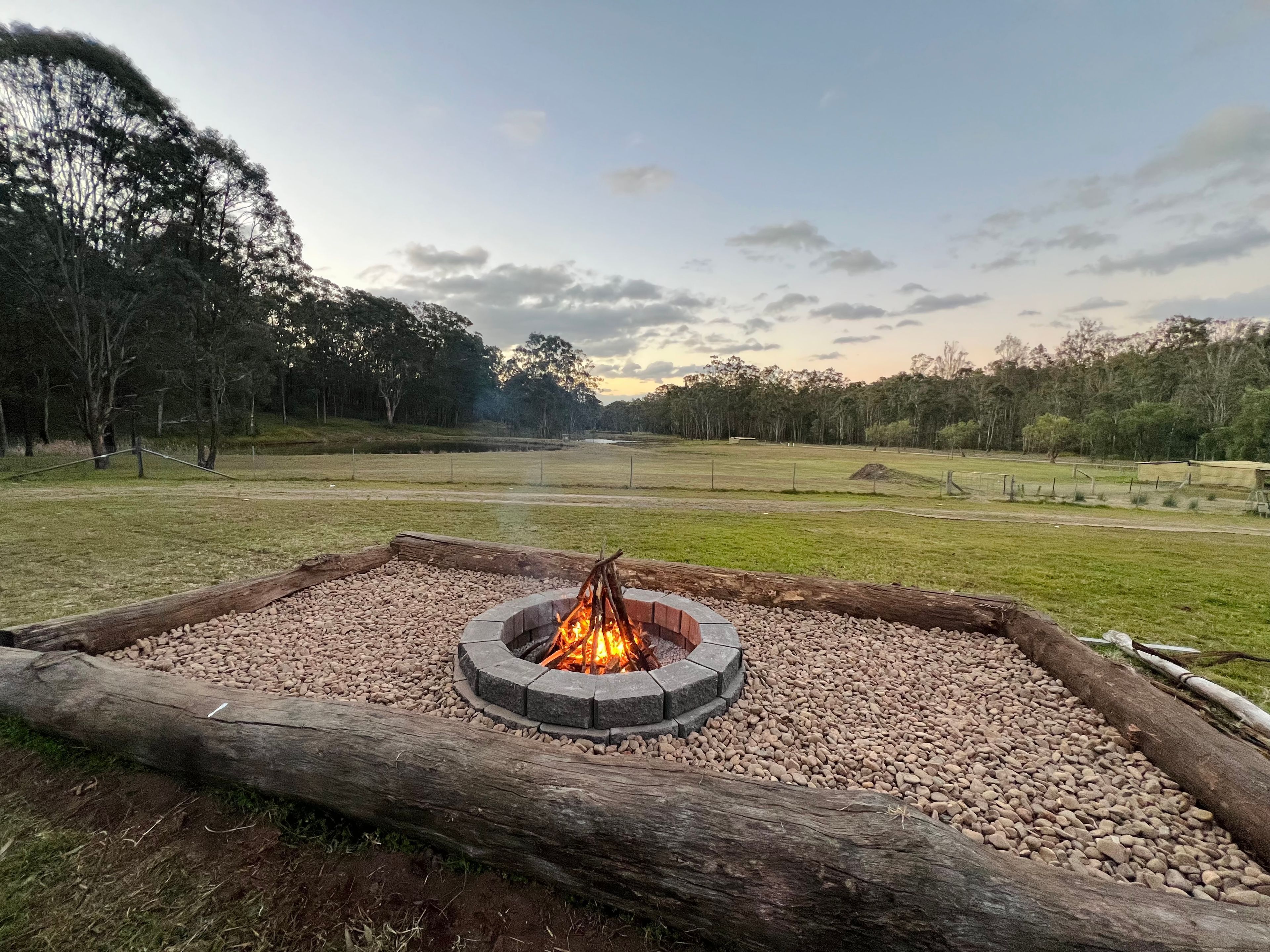 A circular fire pit with a fire burning, surrounded by a gravel bed, set in a grassy field with trees in the background.