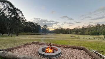 A circular fire pit with a fire burning, surrounded by a gravel bed, set in a grassy field with trees in the background.