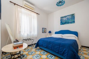 Un dormitorio con cama, una mesa con libros, una ventana con cortinas, un aire acondicionado montado en la pared, una mesita de noche pequeña con una lámpara y un cuadro en la pared.