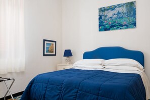 A bedroom with a blue bedspread, a white nightstand with a lamp, a framed picture on the wall, and a painting above the bed. - Casa Simona | Independent Home with Private Garden in the Heart of Positano (Positano)