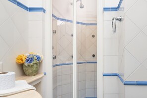 Une salle de bain carrelée de carreaux blancs, une douche avec une porte en verre et une petite étagère sur laquelle est posé un vase de fleurs.